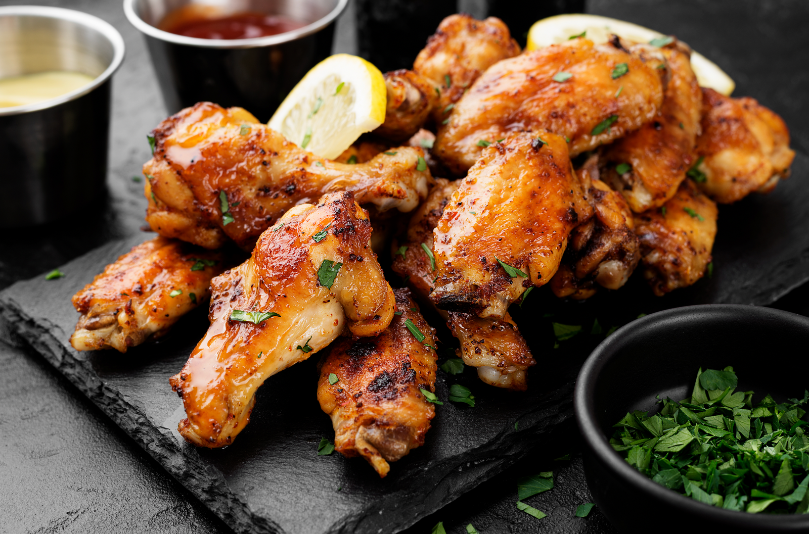 Baked chicken wings on a black slate board with lemon slices and herbs, accompanied by dipping sauces.