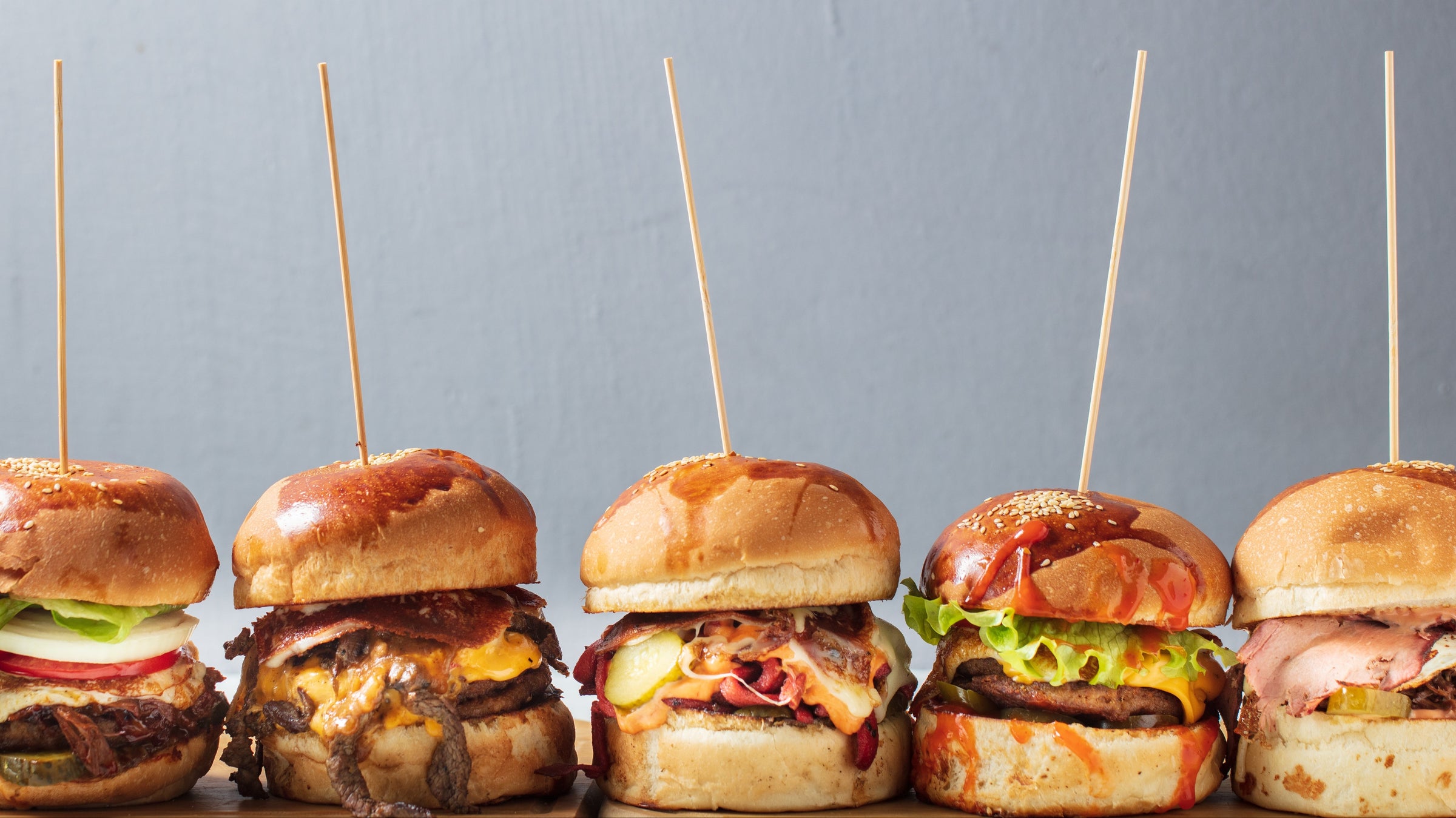 Row of mini burgers with various toppings on a gray background